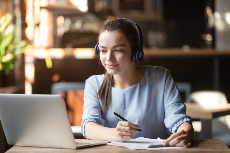 Young woman wearing headphones taking notes while looking at laptop