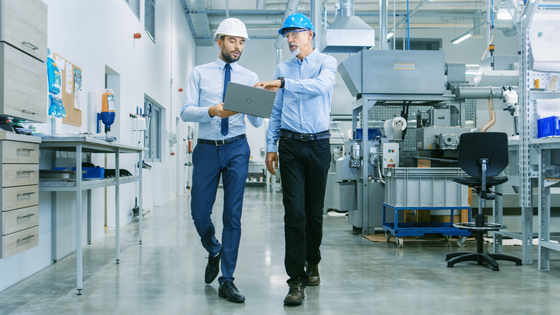 Two workers walking and pointing at laptop one is carrying in industrial space